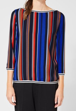 Vrouw draagt een lange mouw top met verticale strepen in blauw, rood, bruin, zwart en wit, gecombineerd met een zwarte broek tegen een effen achtergrond.