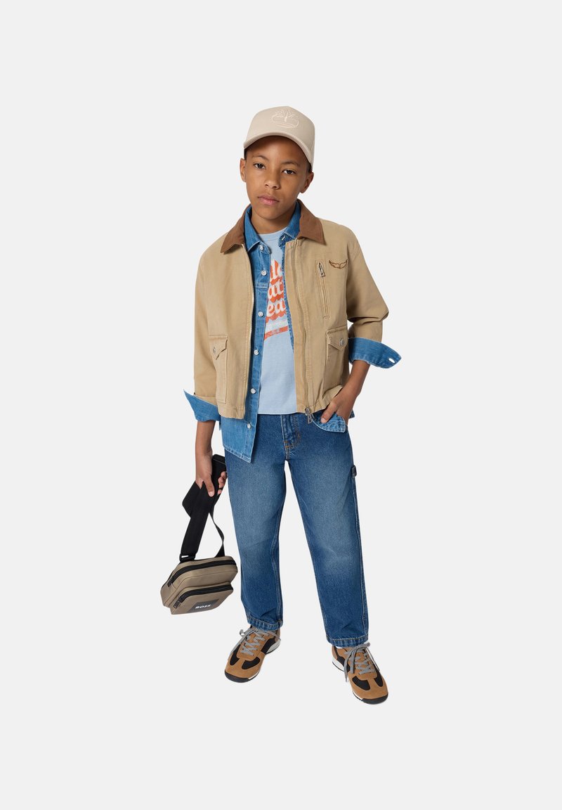 Garçon portant une veste beige sur une chemise en denim bleu et un jean, casquette beige, baskets marron et noires, tenant un sac à bandoulière beige et noir.