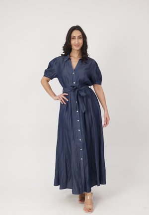 Mujer de pie con la mano en la cadera, vistiendo un vestido maxi azul marino abotonado con mangas abullonadas y cinturón en la cintura, combinado con tacones de tiras transparentes.