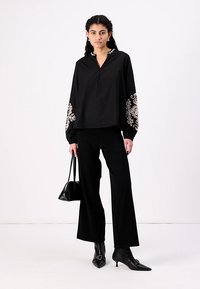 Blusa nera con ricami bianchi sulle maniche, abbinata a pantaloni neri a gamba larga e tacchi neri a punta. Tiene una piccola borsa nera.