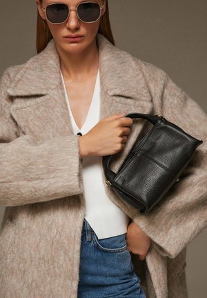 Black textured handbag with a short handle, worn with a beige wool coat over a white top and blue jeans, model wearing round sunglasses.