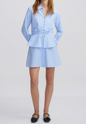 Femme portant un chemisier bleu clair à manches longues boutonné avec une taille évasée et une jupe courte assortie, associée à des chaussures noires.