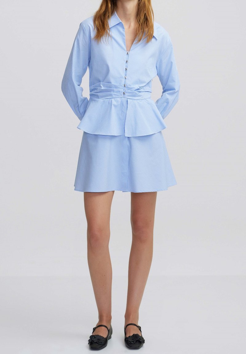 Femme portant un chemisier bleu clair à manches longues boutonné avec une taille évasée et une jupe courte assortie, associée à des chaussures noires.