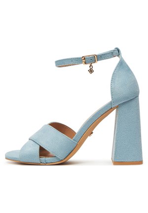 Sandalo con tacco alto in camoscio azzurro chiaro, con ampio cinturino frontale incrociato, cinturino alla caviglia con fibbia dorata e ciondolo, e tacco a blocco.