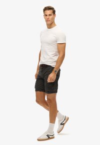 Witte t-shirt met korte mouwen, zwarte cargo shorts met een patch pocket, en witte sneakers met zwarte accenten. Casual outfit met een bruine riem.