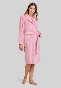 Mujer vestida con un albornoz de rayas rosas hasta la rodilla, con botones y un cinturón atado, de pie descalza frente a un fondo liso.