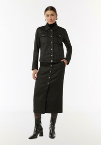 Czarna dżinsowa kurtka z zamknięciem na guziki z przodu oraz kieszeniami naszytymi, zestawiona z pasującą czarną spódnicą dżinsową z rozcięciem z przodu i detalami na guziki.