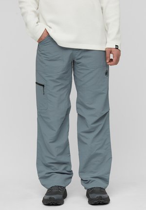 Mann trägt hellblaue Cargohosen mit schwarzer Reißverschlusstasche, weißes strukturiertes Langarmshirt und graue Wanderschuhe, stehend.