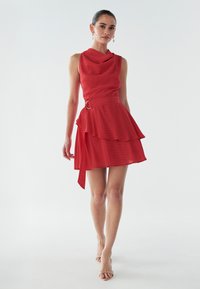 Punainen hihaton mekko, jossa liehuva pääntie, kerroksellinen röyhelöhame, sivutiedolla ja pyöreä solki. Sileä rakenne.