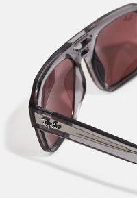 Occhiali da sole Ray-Ban con montatura trasparente grigia, lenti rosse scure e dettagli in argento sulle tempie. Design geometrico con bordi angolari.