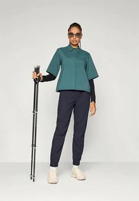 Σκούρο πράσινο μπλουζάκι κοντό μανίκι, μαύρη μακριά κάλτσα από κάτω, ναυτικές tapered παντελόνες και ανοιχτό μπεζ αθλητικά παπούτσια. Μοντέλο holding μαύρους μπαστούνια πεζοπορίας.