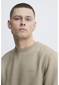 Sudadera beige con cuello redondo y puños acanalados. Presenta un logotipo "blend" en relieve en el pecho. Textura de tela suave, retrato en primer plano.