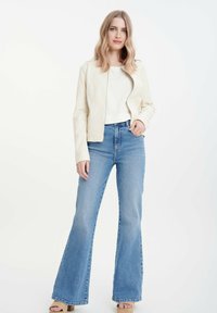 Giacca di pelle attillata color crema con zip, abbinata a jeans a campana blu a vita alta. Il modello si trova su uno sfondo neutro.