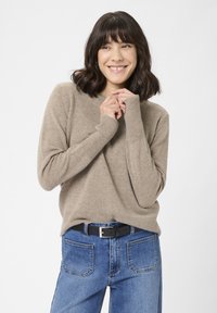 Kvinde med skulderlangt mørkt hår iført lyst sweater og blå jeans, smilende med hænderne foldet nær brystet mod en hvid baggrund.
