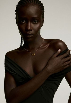 Collarín dorado en forma de media luna sobre una modelo con cabello trenzado; ella lleva un vestido negro off-shoulder que resalta su hombro y escote.