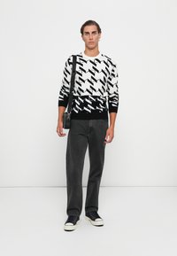 Mann trägt einen schwarz-weiß gemusterten Pullover, eine schwarze Umhängetasche, dunkle Jeans und schwarze Sneaker und steht vor einem weißen Hintergrund.