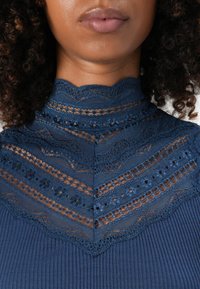 Close-up de uma mulher a usar uma blusa azul de renda com gola alta, apresentando padrões intrincados e tecido canelado abaixo do painel de renda.