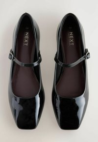 Zapatos Mary Jane de charol negro con puntas cuadradas, una correa delgada en la parte delantera y plantillas acolchadas. Acabado liso y brillante.