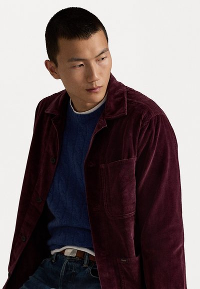 Burgundy corduroy jacket with a notched collar and pocket, layered over a blue cable-knit sweater, paired with dark denim and a belt.