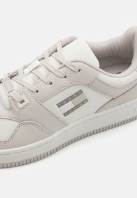 Baskets gris clair avec dessus en cuir texturé, accents en maille blanche, lacets plats et détail de logo métallique sur le côté. Semelle en caoutchouc avec motif antidérapant.