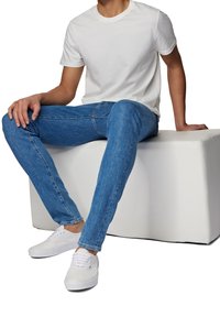 T-shirt en coton blanc, jean en denim bleu et baskets blanches. Le modèle est assis sur un cube blanc, mettant l'accent sur les détails et la coupe des vêtements.