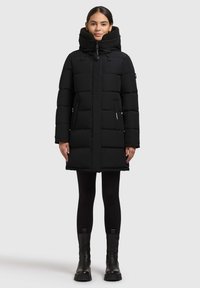 Melnas pūkaino mētelis ar augstu apkakli, kuram ir quilted daļas, stiept akcenti un sānu kabatas, kombinēts ar melniem legingiem un masīviem zābakiem.