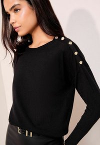 Maglione nero a maglia con scollo rotondo e maniche lunghe, ornato da bottoni dorati lungo la cucitura della spalla. Texture morbida.