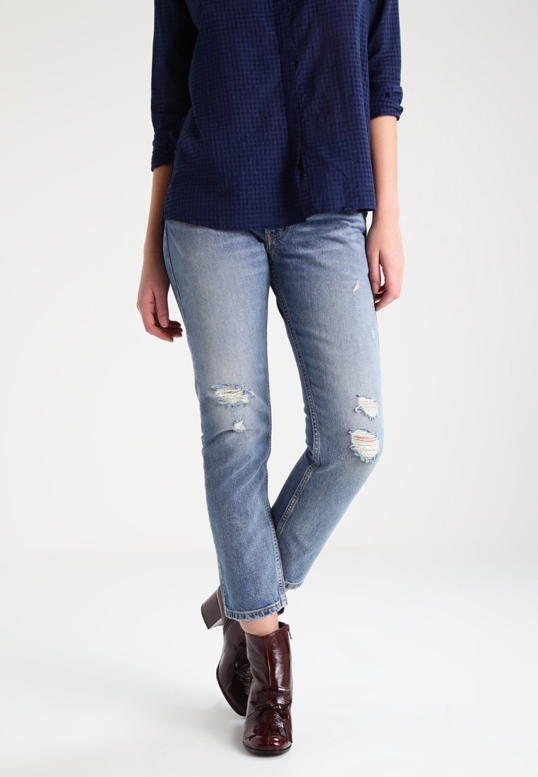 Femme portant un jean déchiré bleu clair, un chemisier à manches longues bleu foncé et des bottines brillantes marron foncé, debout sur un sol blanc.