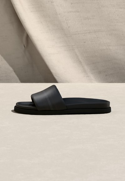 Μαύρη σαγιονάρα με φαρδιά λουρί και εμφανή ραφές, ακουμπισμένη σε φωτεινή επιφάνεια με φόντο μπεζ ύφασμα.