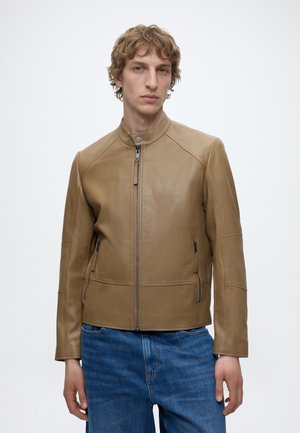 Noor mees, kellel on lühikesed lokkis juuksed, kandmas beežikat nahkjopet ja siniseid teksaseid, seisab tavalise valge tausta ees.