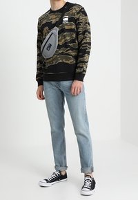 Kampmönstrad svart och grön sweatshirt, ljusblå jeans, svarta låga sneakers och en grå axelväska. Avslappnad outfit.