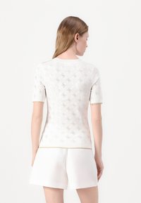 Top bianco a maniche corte con un motivo testurizzato e un logo metallico sottile. Caratterizzato da un colletto rotondo e orlo a contrasto con dettagli dorati.