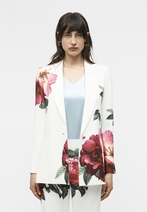 Vrouw draagt een wit pak met grote roze en rode bloemenprints, gecombineerd met een lichtblauwe blouse, staand tegen een effen achtergrond.