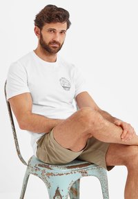 White crew-neck t-shirt featuring a circular graphic on the left chest. Model is seated on a distressed blue metal chair, wearing khaki shorts.