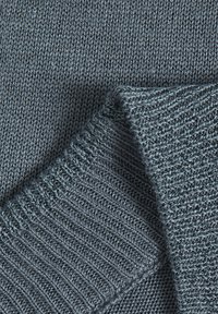 Gros plan sur un tissu tricoté gris plié, montrant les textures détaillées des mailles et les bords à motif côtelé.