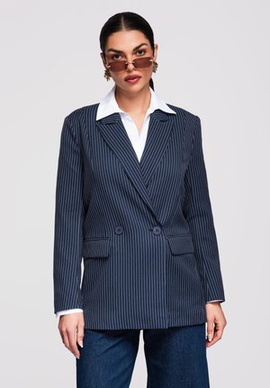 Žena nosící hnědé obdélníkové sluneční brýle, zlaté náušnice, tmavě modrý blazer s proužky, bílou košili a modré džíny na šedém pozadí.