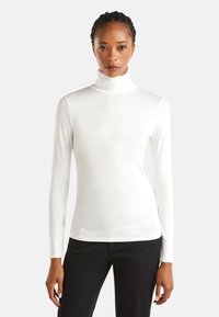Eine Frau trägt einen weißen Rollkragenpullover mit langen Ärmeln und schwarze Hosen. Sie steht vor einem einfachen hellen Hintergrund und blickt geradeaus.