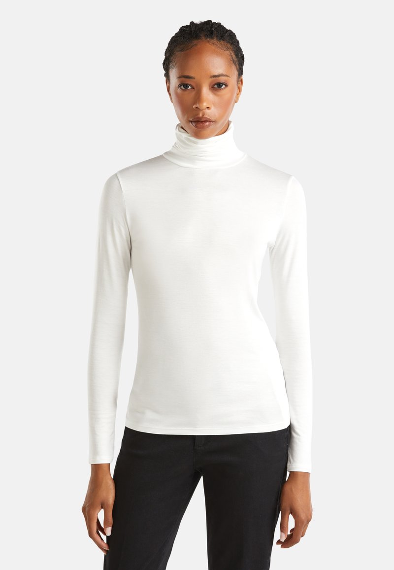 Eine Frau trägt einen weißen Rollkragenpullover mit langen Ärmeln und schwarze Hosen. Sie steht vor einem einfachen hellen Hintergrund und blickt geradeaus.