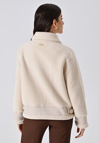 Giacca in pile color crema con una consistenza morbida, vestibilità rilassata e accenti beige a contrasto. Presenta un colletto e un piccolo dettaglio logo dorato.