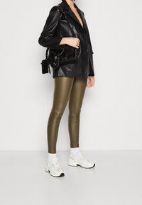 Svart läderblazer, olivgröna leggings, vita strumpor och ljusa sneakers. Handväska med texturerade detaljer hänger från axeln.