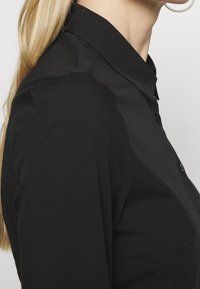 Zwart overhemd met kraag en knopen, met een gladde textuur en aansluitend ontwerp, vanaf de zijkant weergegeven tegen een neutrale achtergrond.