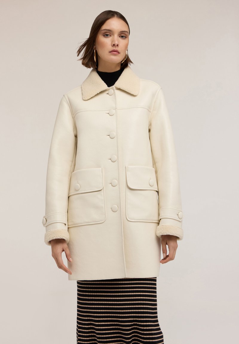 Cappotto in pelle color crema con chiusura a bottoni, colletto oversize e due tasche frontali. Polsini e colletto foderati in shearling per una texture aggiuntiva.