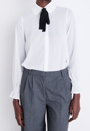 Personne portant un chemisier blanc à manches longues avec un col nœud papillon noir et un pantalon plissé taille haute gris sur un fond uni.