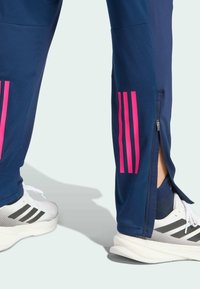 Lower legs wearing navy blue athletic pants with pink stripes and side zippers, paired with white sneakers featuring black stripes.