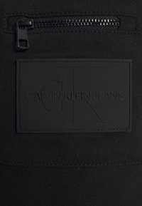 Zwarte stof met een zwart rubberen Calvin Klein Jeans logo patch en een zwarte ritszak boven de patch.