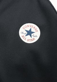 Στρογγυλό λευκό σήμα Converse All Star με μπλε αστέρι και κόκκινα γράμματα σε μαύρο υφασμάτινο φόντο με υφή.