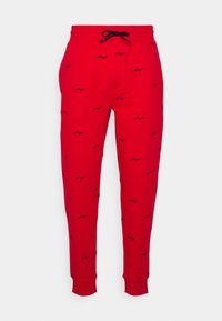 Rote Jogginghose mit Kordelzugbund, einem sich wiederholenden schwarzen Logomuster, zulaufenden Beinen und gerippten Bündchen an den Knöcheln.