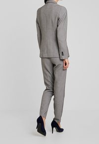 Grau karierter Anzug mit tailliertem Blazer und schmalen Hosen. Hat eine glatte Textur und schwarze Knopfakzente, kombiniert mit marineblauen High Heels.