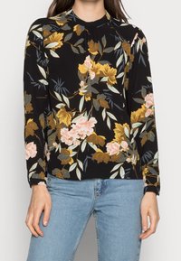 Blouse fleurie sur fond noir, ornée de fleurs jaunes, roses et vertes. Manches longues avec un col arrondi et une texture de tissu délicate.