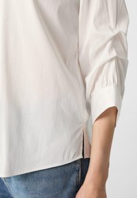 Hellbeige Bluse mit lockerer Passform, gerafften Ärmeln und seitlichen Schlitzen, aus einem glatten Stoff gefertigt. Kombiniert mit Jeans.